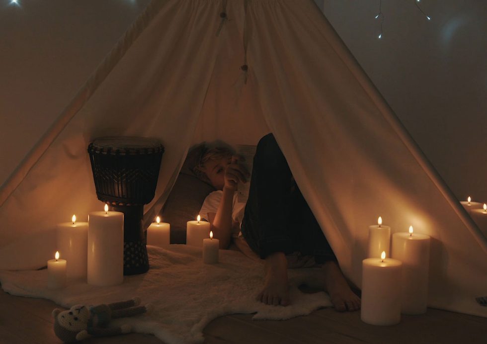 Child reading in a cosy teepee surrounded by Uyuni Lighting Classic Ivory Flameless Pillar Candles, creating a safe, warm and inviting children’s room.
